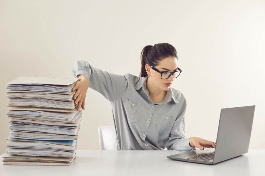 Women on computer at a desk beside a pile of paper documents 